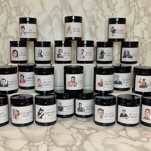 May include: A collection of 24 candles with black glass jars and labels featuring portraits of various celebrities. Each candle has a different celebrity's name and a short description.