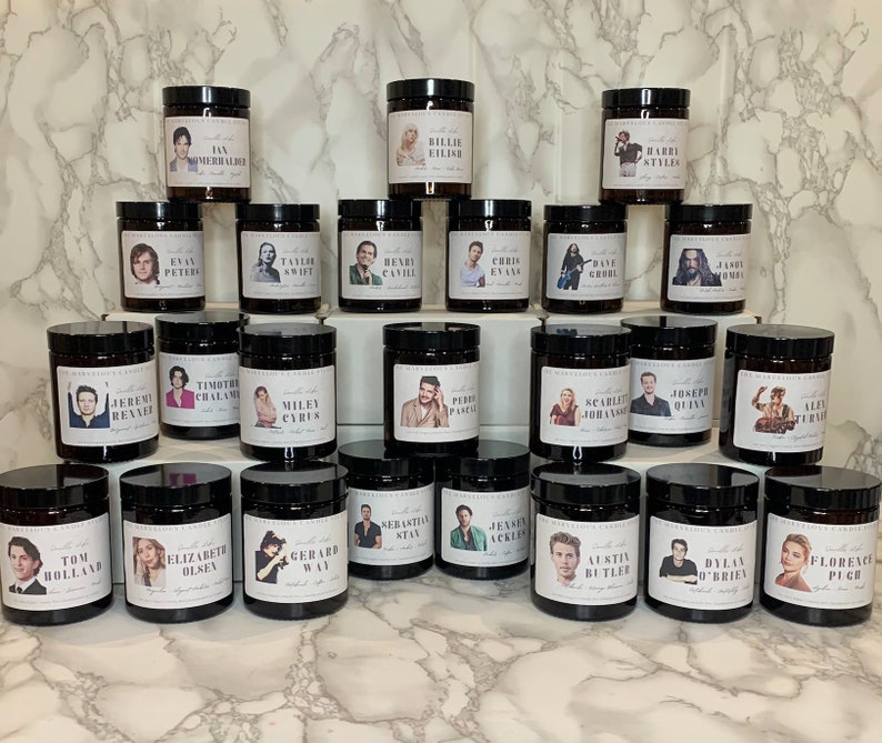 May include: A collection of 24 candles with black glass jars and labels featuring portraits of various celebrities. Each candle has a different celebrity's name and a short description.