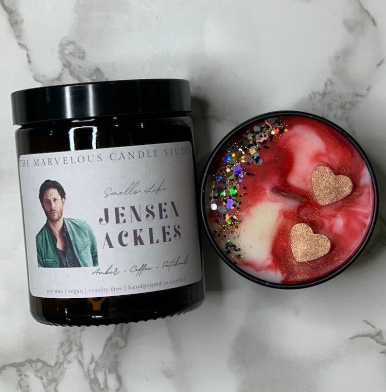 May include: A soy wax candle with a black lid and a label that reads "The Marvelous Candle Studio" and "Smells Like Jensen Ackles". The candle is decorated with glitter and two gold hearts. The label also states "Amber - Coffee - Patchouli" and "Soy wax | Vegan | Cruelty free | Handpoured in London."