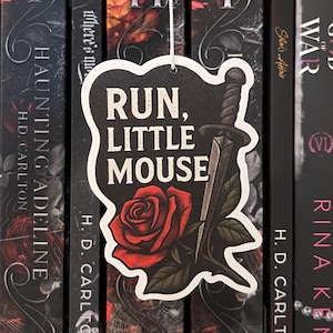 Peut inclure: Marque-page noir et blanc avec le texte "RUN, LITTLE MOUSE" et une illustration de rose et de poignard. Le marque-page est suspendu devant une rangée de livres aux couvertures sombres.