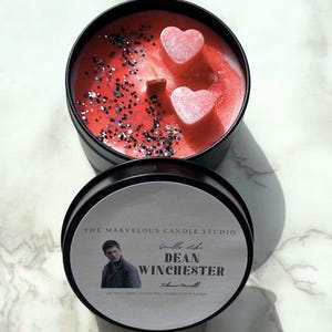 Op de afbeelding: Een zwarte kaarsenblik met een rode en zilveren glitterkaars erin. De kaars heeft twee roze hartvormige wasdecoraties bovenop. Het etiket op het blik zegt "Smell Like Dean Winchester" met een foto van Jensen Ackles. De tekst op het etiket zegt ook "The Marvelous Candle Studio" en "Handmade in London."