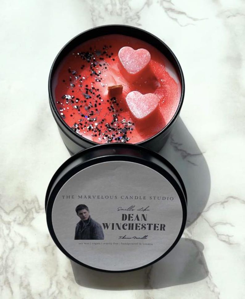 Op de afbeelding: Een zwarte kaarsenblik met een rode en zilveren glitterkaars erin. De kaars heeft twee roze hartvormige wasdecoraties bovenop. Het etiket op het blik zegt "Smell Like Dean Winchester" met een foto van Jensen Ackles. De tekst op het etiket zegt ook "The Marvelous Candle Studio" en "Handmade in London."