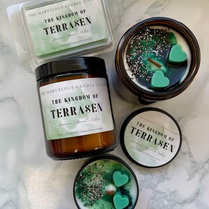 May include: An assortment of scented candles and wax melts. The candles are in brown glass jars and tins, with green wax and heart-shaped accents. The wax melts are in a clear plastic container. The labels read "THE KINGDOM OF TERRASEN".