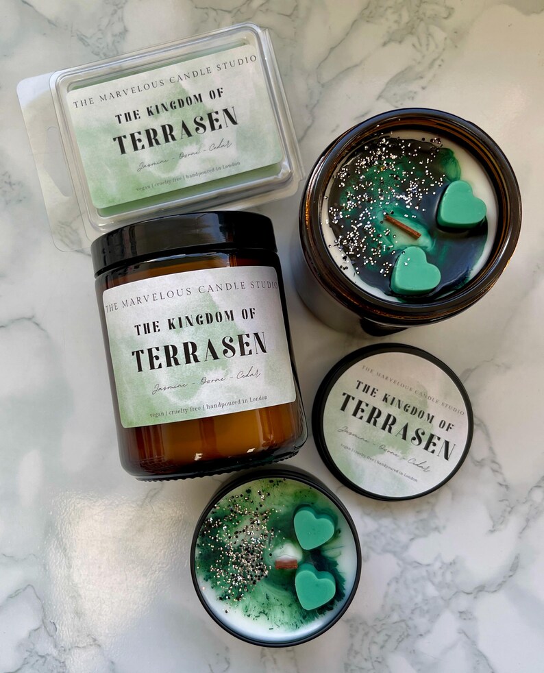 May include: An assortment of scented candles and wax melts. The candles are in brown glass jars and tins, with green wax and heart-shaped accents. The wax melts are in a clear plastic container. The labels read "THE KINGDOM OF TERRASEN".