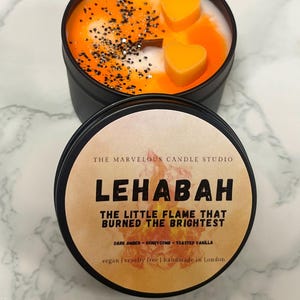 May include: A black candle tin with a lid, featuring a candle with orange and white wax, orange heart-shaped wax melts, and black glitter. The label reads "LEHABAH THE LITTLE FLAME THAT BURNED THE BRIGHTEST".