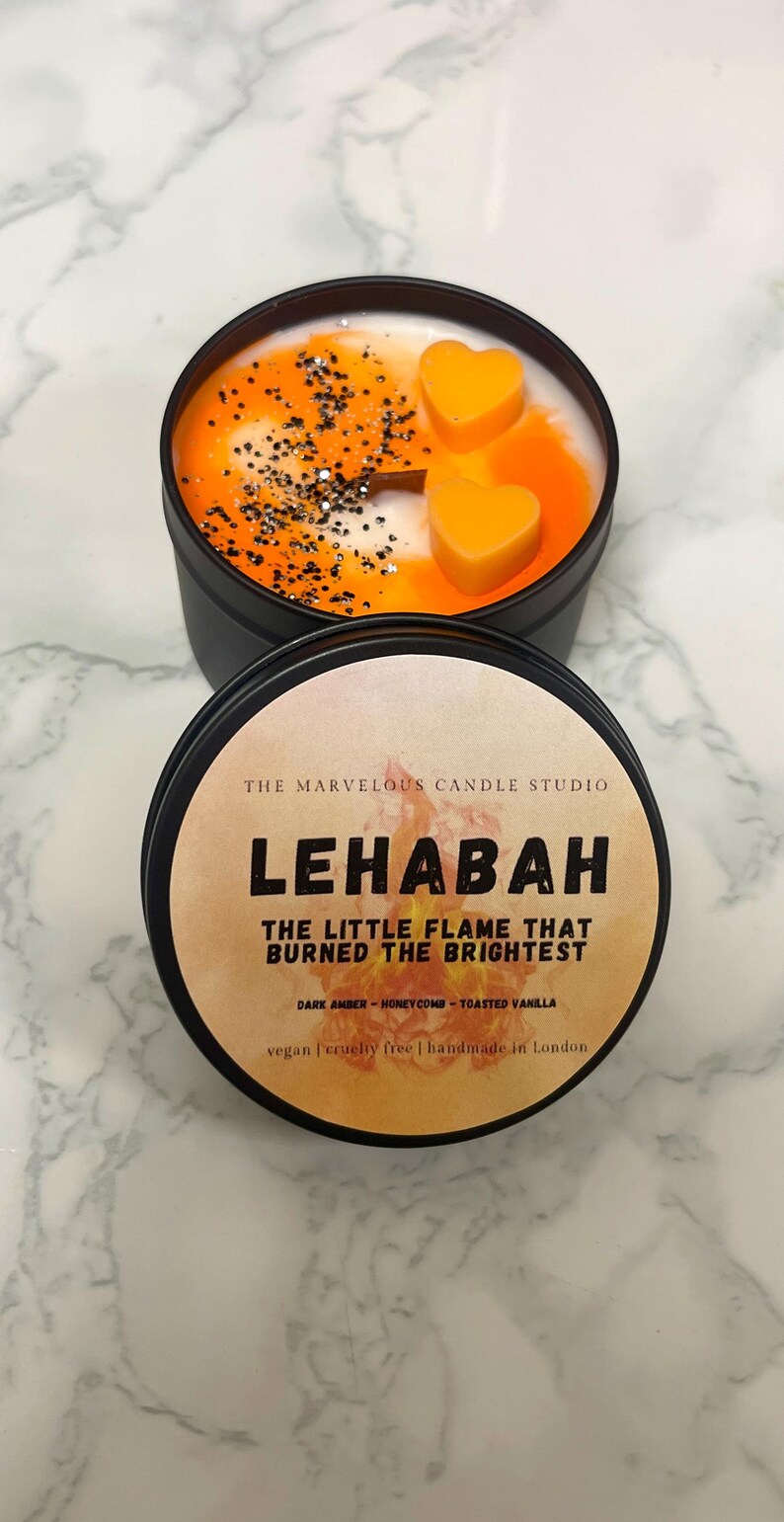May include: A black candle tin with a lid, featuring a candle with orange and white wax, orange heart-shaped wax melts, and black glitter. The label reads "LEHABAH THE LITTLE FLAME THAT BURNED THE BRIGHTEST".