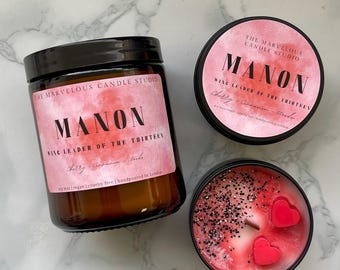May include: Three candles from The Marvelous Candle Studio. One is in a glass jar with a black lid, and two are in black tins. All have pink labels with the name "MANON" and "WING LEADER OF THE THIRTEEN". The open candle has red heart-shaped wax accents.