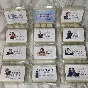K&ouml;nnte beinhalten: Zw&ouml;lf wei&szlig;e quadratische Kerzenbeh&auml;lter mit durchsichtigen Plastikdeckeln. Jeder Beh&auml;lter hat ein anderes Foto und den Namen einer Ber&uuml;hmtheit auf der Vorderseite gedruckt. Der Text "The Marvelous Candle Studio" ist oben auf jedem Beh&auml;lter gedruckt.