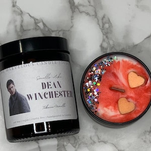 Op de afbeelding: Een zwarte kaars pot met een label dat "The Marvelous Candle Studio" en "Smells Like Dean Winchester" zegt met een foto van een man. De kaars is van sojawas, veganistisch, wreedheidvrij en handgemaakt in Londen. De kaars is tabak-vanille geur. De kaars zit in een zwarte pot met een rood en wit swirl ontwerp en twee perzikkleurige harten.