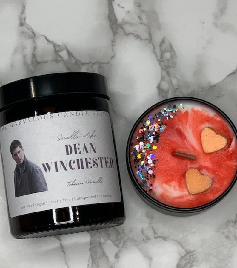 Op de afbeelding: Een zwarte kaars pot met een label dat "The Marvelous Candle Studio" en "Smells Like Dean Winchester" zegt met een foto van een man. De kaars is van sojawas, veganistisch, wreedheidvrij en handgemaakt in Londen. De kaars is tabak-vanille geur. De kaars zit in een zwarte pot met een rood en wit swirl ontwerp en twee perzikkleurige harten.