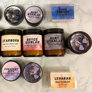 May include: An assortment of scented candles and wax melts. The candles are in glass jars and metal tins, with labels featuring text like "Hunt Athalar" and "Starborn." The wax melts are in clear plastic containers. Colors include purple, blue, and gold.
