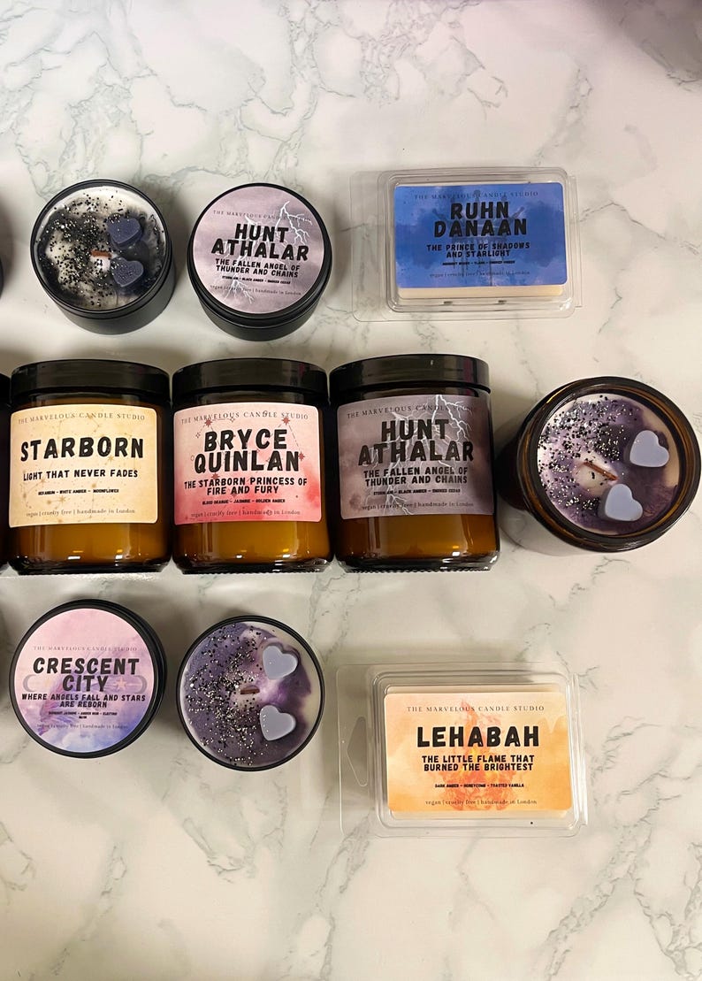 May include: An assortment of scented candles and wax melts. The candles are in glass jars and metal tins, with labels featuring text like "Hunt Athalar" and "Starborn." The wax melts are in clear plastic containers. Colors include purple, blue, and gold.