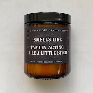 Puede incluir: Una vela en tarro de cristal con tapa negra. La etiqueta dice "SMELLS LIKE TAMLIN ACTING LIKE A LITTLE BITCH" con "THE MARVELOUS CANDLE STUDIO" en la parte superior. La vela es de cera de soja, vegana y hecha a mano en Londres.