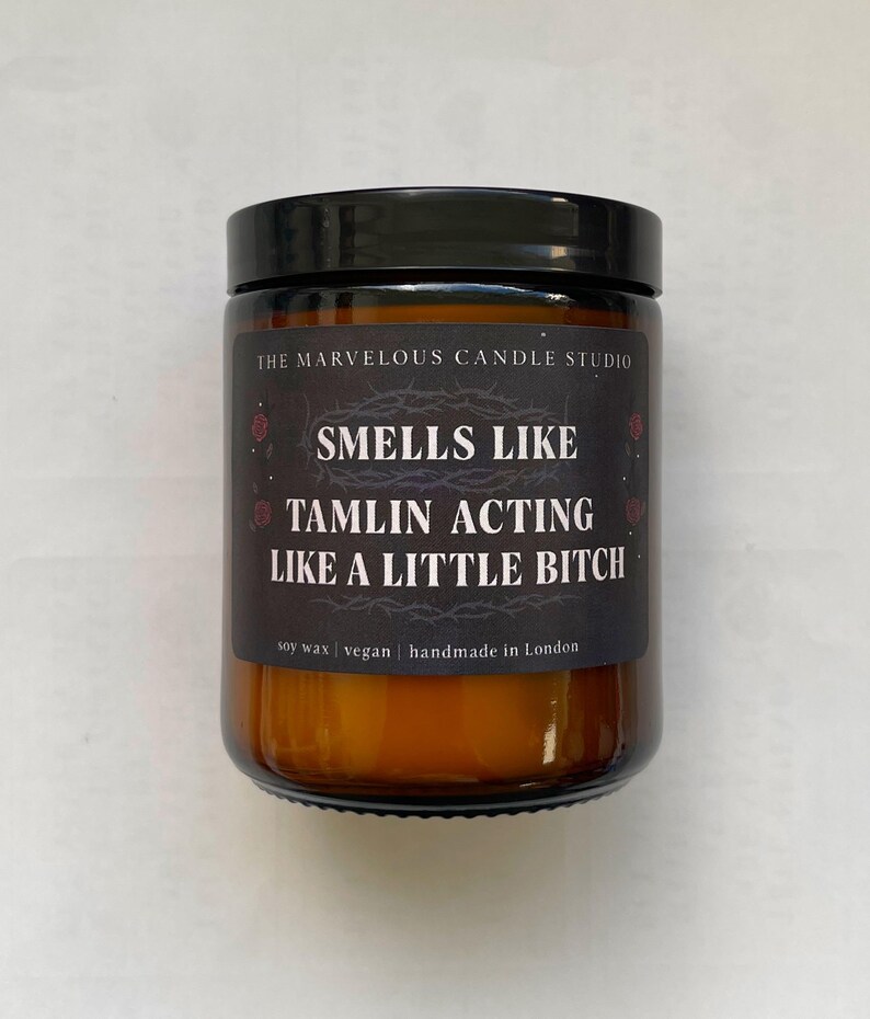 Puede incluir: Una vela en tarro de cristal con tapa negra. La etiqueta dice "SMELLS LIKE TAMLIN ACTING LIKE A LITTLE BITCH" con "THE MARVELOUS CANDLE STUDIO" en la parte superior. La vela es de cera de soja, vegana y hecha a mano en Londres.