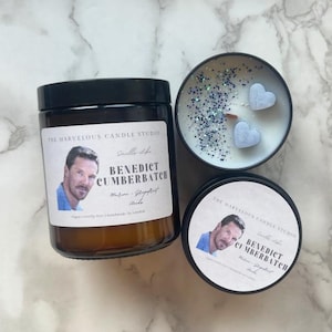 May include: Two candles with black lids and labels featuring a man's face and the text "The Marvelous Candle Studio" and "Benedict Cumberbatch". The candles are on a white marble surface. One candle has a clear lid with blue glitter and two blue heart-shaped decorations.