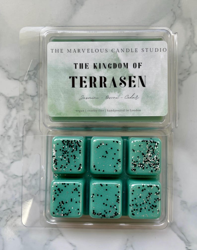 May include: A clear plastic clamshell package containing six teal wax melts. Each melt is square and speckled with black and silver glitter. The label reads "THE KINGDOM OF TERRASEN" and lists the scent as Jasmine, Derne, and Cedar.