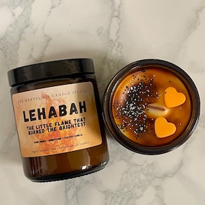 May include: Two brown glass jar candles with black lids. One candle has a label that reads "LEHABAH" and "THE LITTLE FLAME THAT BURNED THE BRIGHTEST." The other candle has two orange heart-shaped wax accents. The candles are on a white marble surface.
