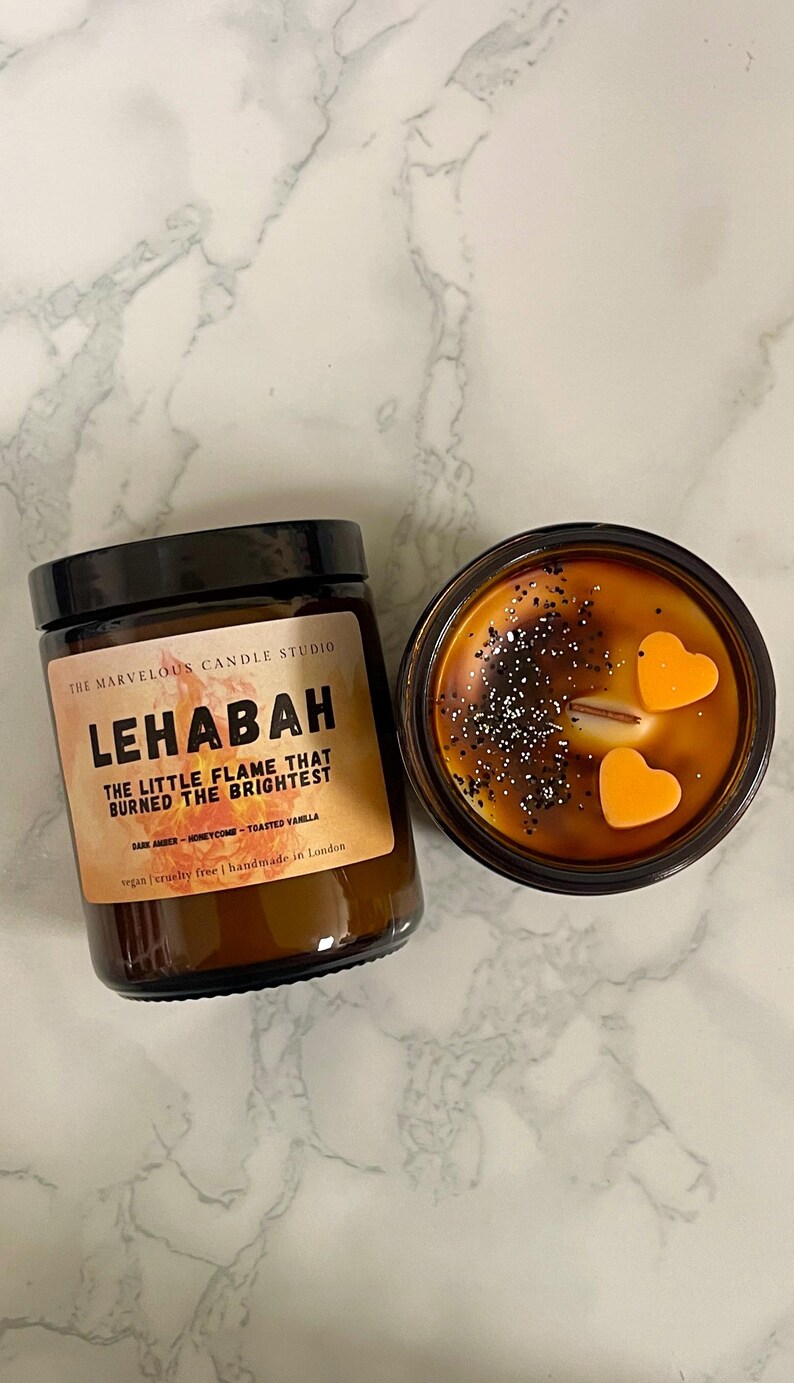May include: Two brown glass jar candles with black lids. One candle has a label that reads "LEHABAH" and "THE LITTLE FLAME THAT BURNED THE BRIGHTEST." The other candle has two orange heart-shaped wax accents. The candles are on a white marble surface.