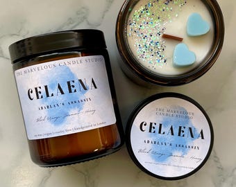 May include: Three brown glass jar candles with black lids. The labels read "CELAENA" and "ADARIAN'S ASSASSIN". One candle has blue heart-shaped wax melts and glitter. The text on the label says "Blood Orange Jasmine - Honey".
