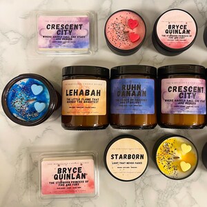 May include: An assortment of scented candles and wax melts from The Marvelous Candle Studio. The candles are in black tins and glass jars, with labels featuring names like "Crescent City" and "Bryce Quinlan." Some candles have heart-shaped wax accents.