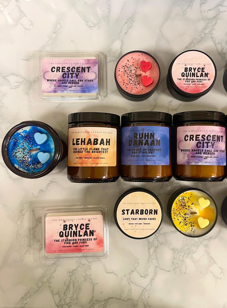 May include: An assortment of scented candles and wax melts from The Marvelous Candle Studio. The candles are in black tins and glass jars, with labels featuring names like "Crescent City" and "Bryce Quinlan." Some candles have heart-shaped wax accents.