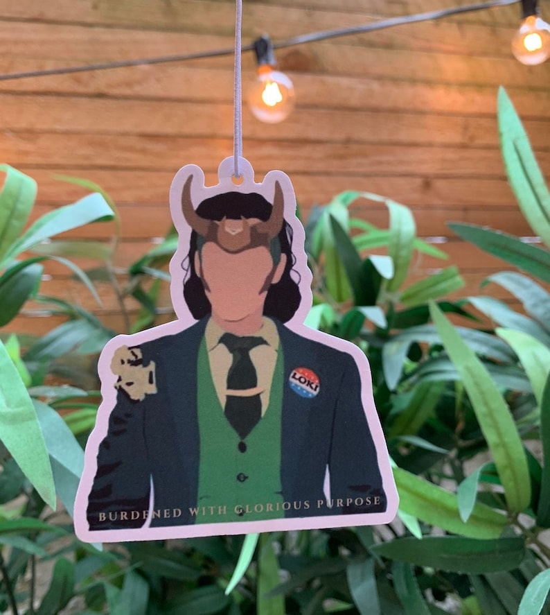 May include: A white car air freshener featuring a cartoon illustration of Loki from the Marvel Cinematic Universe. He is wearing a green suit, a green vest, and a gold crown. The text "BURDENED WITH GLORIOUS PURPOSE" is printed below the image. The text "LOKI" is printed on a blue button on his suit.