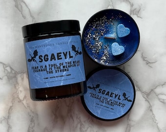 May include: Two candles with a blue and silver design. The candles have a black label with the text "SGAEYL FEAR IS A TOOL OF THE WEAK. COURAGE IS THE WEAPON OF THE STRONG." One candle is in a glass jar and the other is in a metal tin.