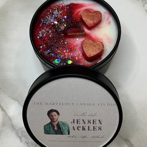 May include: A black candle tin with a white label featuring a photo of Jensen Ackles. The label also includes the text "Smells Life", "Jensen Ackles", "Amber - Coffee - Patchouli", "Soy Wax | Vegan | Cruelty Free | Handpoured in London". The candle tin has a red and white wax design with two red glitter hearts.