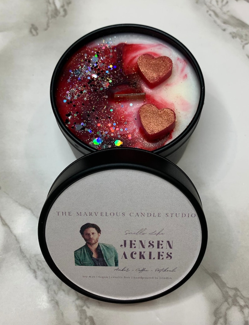 May include: A black candle tin with a white label featuring a photo of Jensen Ackles. The label also includes the text "Smells Life", "Jensen Ackles", "Amber - Coffee - Patchouli", "Soy Wax | Vegan | Cruelty Free | Handpoured in London". The candle tin has a red and white wax design with two red glitter hearts.
