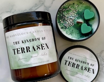 May include: An assortment of scented candles from The Marvelous Candle Studio. One candle is in a brown glass jar with a black lid, and the label reads "The Kingdom of Terrasen." Another candle is in a black tin with green wax and heart-shaped accents.