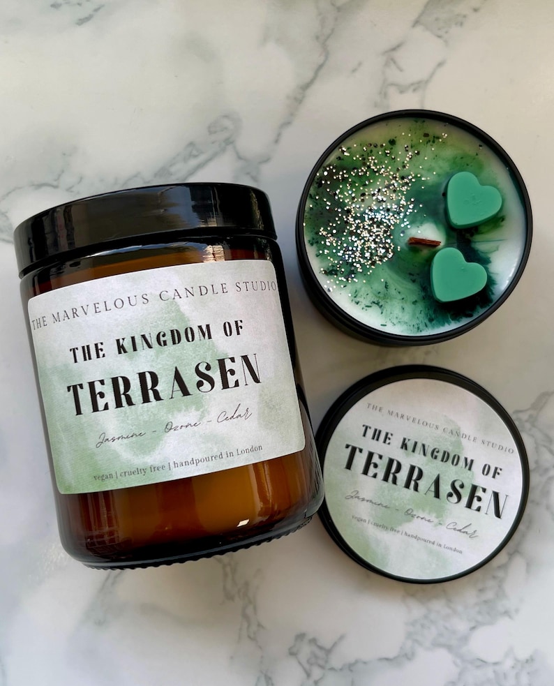 May include: An assortment of scented candles from The Marvelous Candle Studio. One candle is in a brown glass jar with a black lid, and the label reads "The Kingdom of Terrasen." Another candle is in a black tin with green wax and heart-shaped accents.