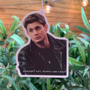 Puede incluir: Un ambientador de coche blanco con una foto de un hombre con cabello castaño y una chaqueta de cuero negra. El texto "Demons I get, people are crazy" está impreso debajo de la imagen.