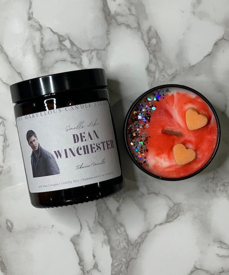 Op de afbeelding: Een rode en witte kaars met een zwarte deksel en een label met de tekst "Smells Like Dean Winchester Tobacco Vanille" en een foto van Dean Winchester. De kaars is omgeven door glitter en heeft twee hartvormige decoraties bovenop.