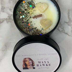 K&ouml;nnte beinhalten: Ein schwarzes Kerzengef&auml;&szlig; mit einem wei&szlig;en Etikett, auf dem "The Marvelous Candle Studio" und "Maya Hawke" mit dem Duft "Lychee & Peony" steht. Das Gef&auml;&szlig; ist mit wei&szlig;em Wachs gef&uuml;llt und mit goldenem Glitzer und zwei wei&szlig;en herzf&ouml;rmigen Wachsdekorationen verziert.