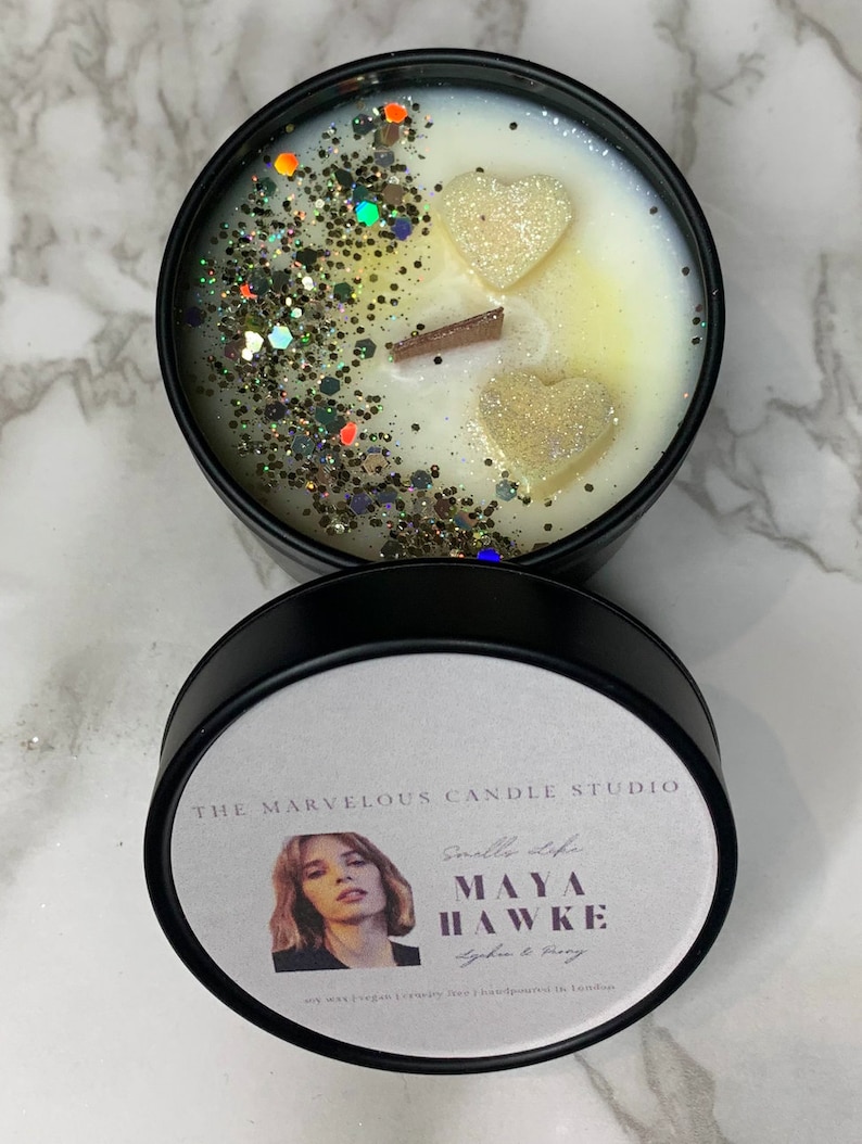 K&ouml;nnte beinhalten: Ein schwarzes Kerzengef&auml;&szlig; mit einem wei&szlig;en Etikett, auf dem "The Marvelous Candle Studio" und "Maya Hawke" mit dem Duft "Lychee & Peony" steht. Das Gef&auml;&szlig; ist mit wei&szlig;em Wachs gef&uuml;llt und mit goldenem Glitzer und zwei wei&szlig;en herzf&ouml;rmigen Wachsdekorationen verziert.