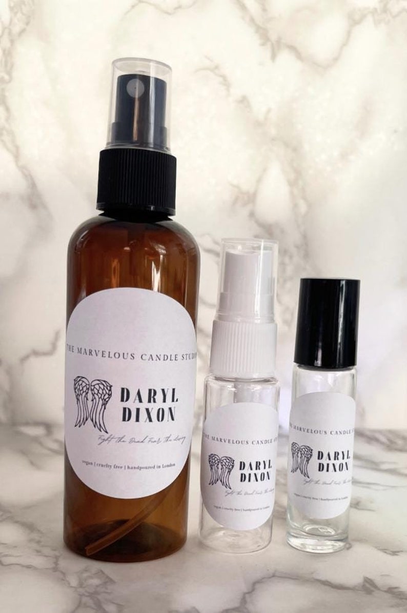 May include: Three glass bottles with black caps and white labels. The labels feature a logo with wings and the text "Daryl Dixon" and "The Marvelous Candle Studio". The text on the label also says "Fight the Bad, Fuel the Story" and "Vegan Cruelty Free | Handpoured in London".