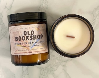 May include: Two brown glass jar candles. One candle has a label that reads "OLD BOOKSHOP WHERE STORIES NEVER SLEEP". The other candle has a white wax with a wooden wick. The candles are on a marble surface.
