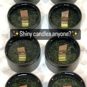 May include: Several black tins filled with a green, glittery substance and a wooden wick. The text "Shiny candles anyone?" is displayed on the image. The candles appear to be handmade.