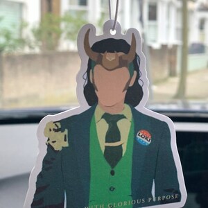 May include: A white car air freshener featuring a cartoon illustration of Loki from the Marvel Cinematic Universe. He is wearing a green suit, a green vest, and a brown horned helmet. The text "Burdened with Glorious Purpose" is printed at the bottom of the air freshener.
