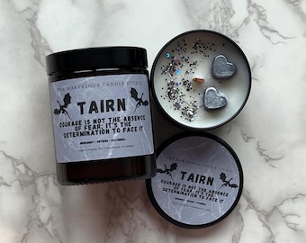 May include: Two black candle jars with a white label featuring a dragon graphic and the text "Tairn: Courage is not the absence of fear: It's the determination to face it." The candle jars are on a white marble surface. One jar has a lid and the other is open, revealing a white candle with silver glitter and two silver heart-shaped charms.