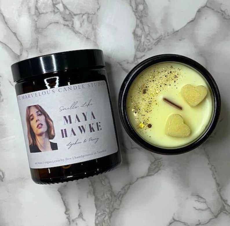 K&ouml;nnte beinhalten: Ein schwarzes Glasgef&auml;&szlig; mit einer Kerze und einem wei&szlig;en Etikett mit der Aufschrift "Smells Life MAYA HAWKE Lychee & Peony" und "Soy wax | Vegan | Cruelty free | Handpoured in London". Die Kerze ist mit gelbem Wachs und zwei herzf&ouml;rmigen Wachsdekorationen verziert.