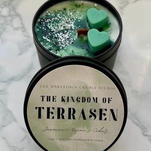 May include: A black candle tin with a green and white wax interior, featuring two heart-shaped teal wax melts and silver glitter. The label reads "THE KINGDOM OF TERRASEN" with scent notes and production details.