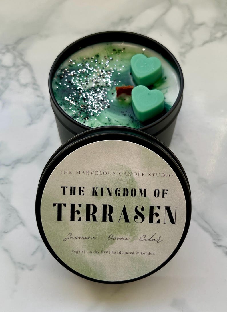 May include: A black candle tin with a green and white wax interior, featuring two heart-shaped teal wax melts and silver glitter. The label reads "THE KINGDOM OF TERRASEN" with scent notes and production details.