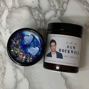 May include: A soy wax candle with a blue and silver glitter design. The candle is in a glass jar with a black lid. The label on the jar says "The Marvelous Candle Studio" and "Smells Like Sam Rockwell".