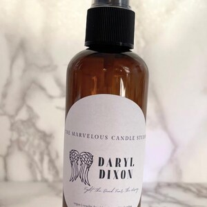 May include: A brown glass bottle with a black spray top and a white label. The label has a black and white illustration of angel wings and the text "Daryl Dixon" and "The Good, The Bad, The Ugly".
