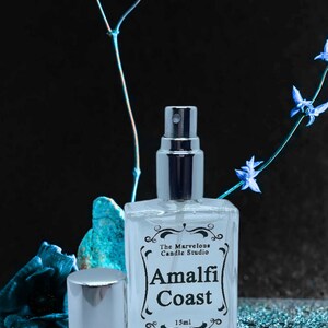 Puede incluir: Una botella de vidrio transparente con una boquilla de pulverizaci&oacute;n plateada y una tapa. La botella est&aacute; etiquetada con "The Marvelous Candle Studio Amalfi Coast 15ml". La botella est&aacute; sobre una superficie brillante azul y plateada.