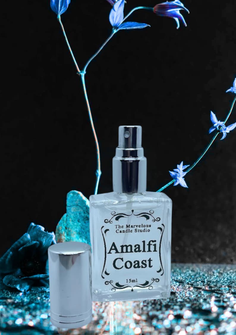 Puede incluir: Una botella de vidrio transparente con una boquilla de pulverizaci&oacute;n plateada y una tapa. La botella est&aacute; etiquetada con "The Marvelous Candle Studio Amalfi Coast 15ml". La botella est&aacute; sobre una superficie brillante azul y plateada.