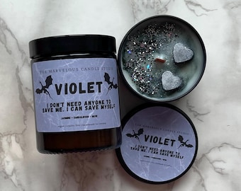 May include: Three candles are displayed on a marble surface. One is a glass jar candle with a black lid, labeled "VIOLET." The candle's label includes text and dragon illustrations. The other two are in black tins, one open, revealing a candle with silver glitter and heart-shaped accents.