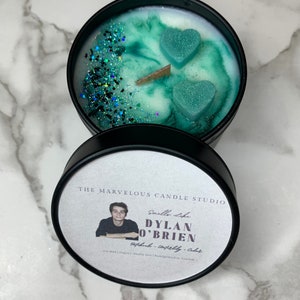 May include: A black candle jar with a white label that reads "The Marvelous Candle Studio" and "Smells Like Dylan O'Brien" with a photo of a man. The candle jar has a green and white wax with two teal heart-shaped wax pieces on top.