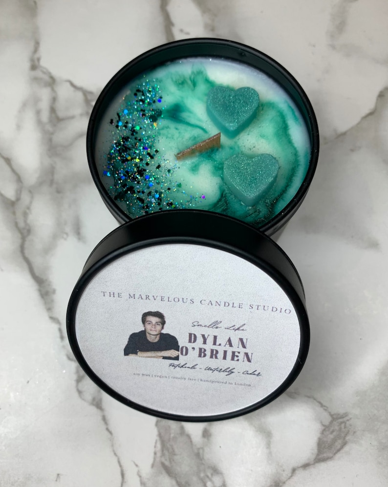 May include: A black candle jar with a white label that reads "The Marvelous Candle Studio" and "Smells Like Dylan O'Brien" with a photo of a man. The candle jar has a green and white wax with two teal heart-shaped wax pieces on top.